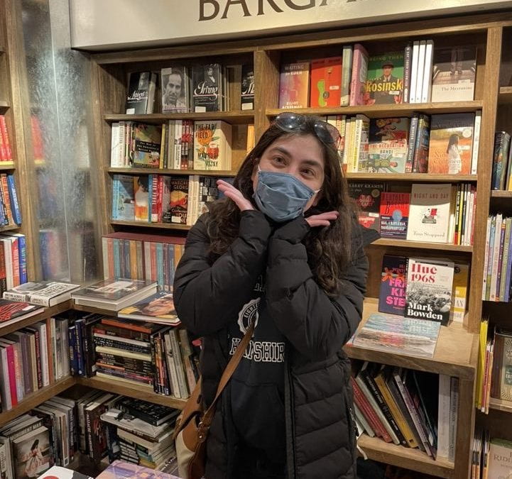 Rachel in a bookstore. She's wearing a mask and a black coat. She is surrounded by shelves of books.