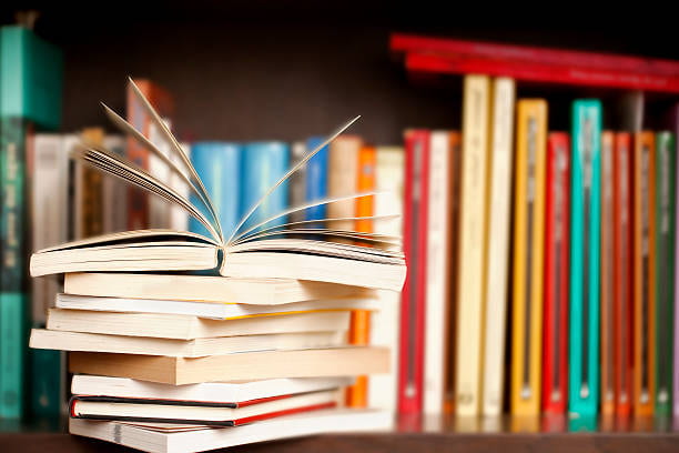 stock photo of a haphazard stack of books. the top book is open. in the background is a shelf of more books, standing.